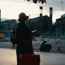 woman wearing coat standing on road with travel luggage during daytime