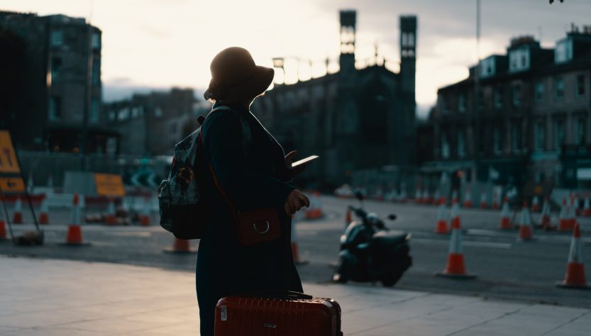 woman wearing coat standing on road with travel luggage during daytime